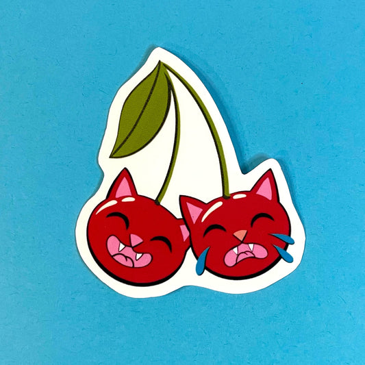 Two cherries shaped like cat heads, bright red and green. One cat is smiling, the other is crying.