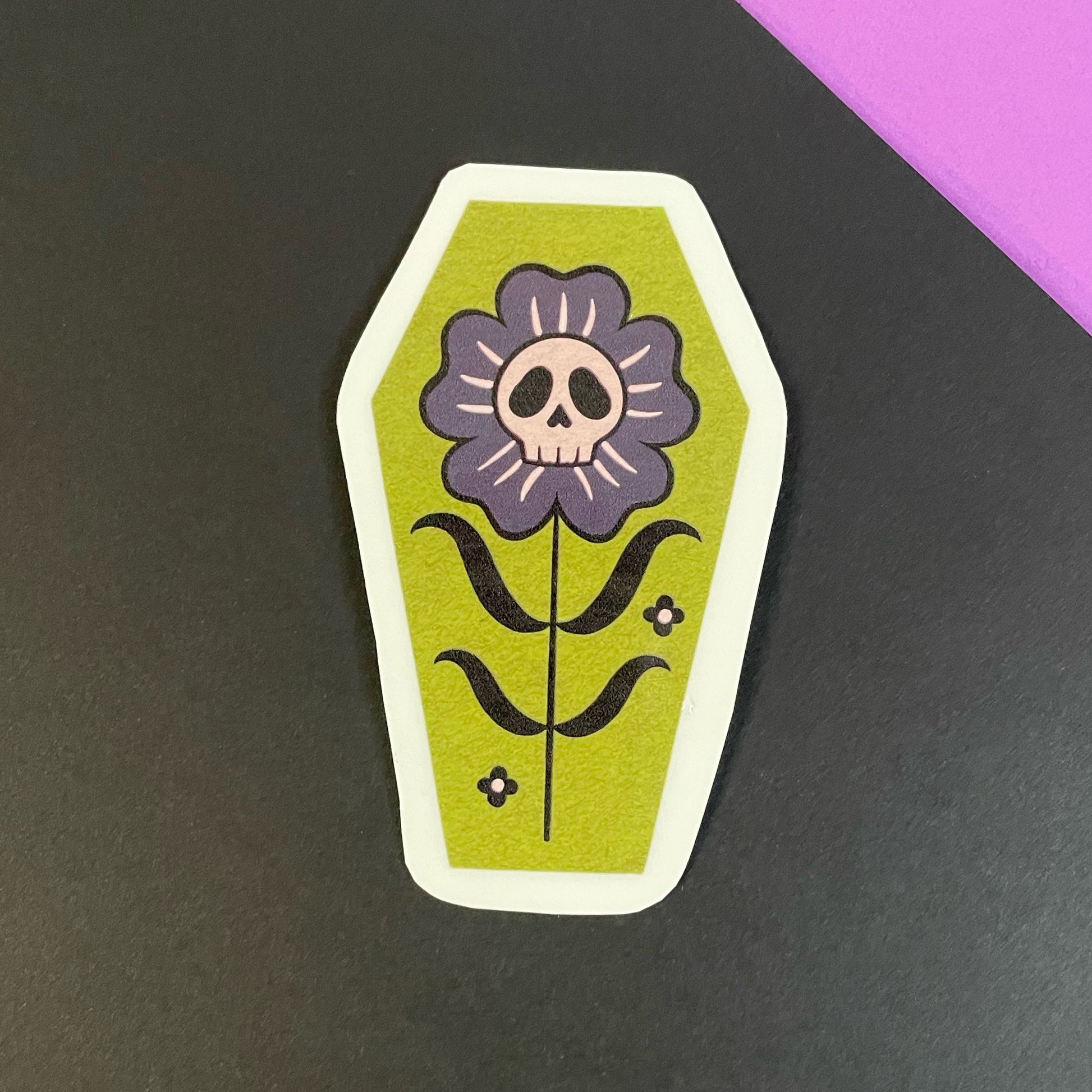 A coffin shaped sticker with a green background, featuring a black and gray flower with a skull in the center.