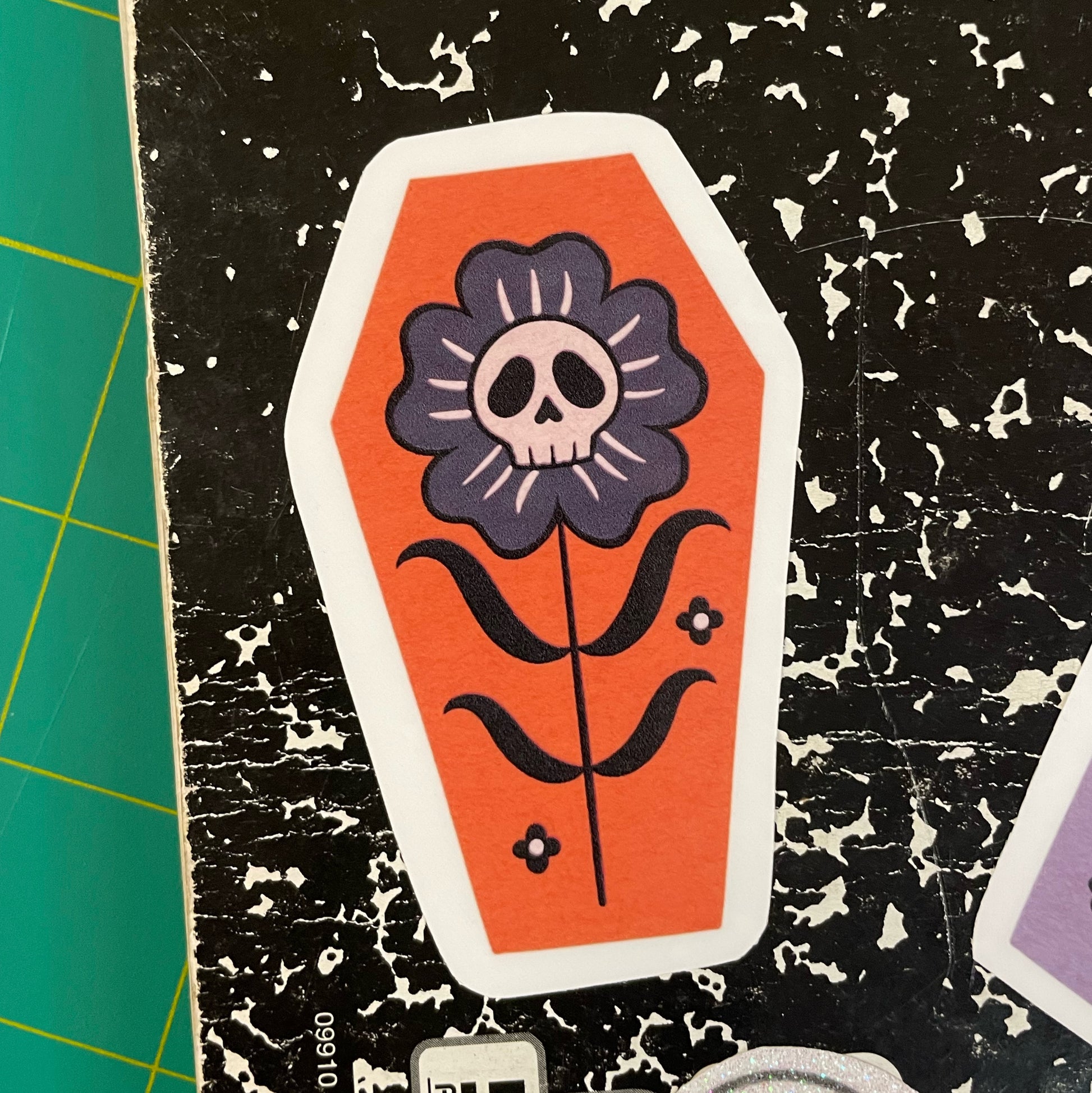 A coffin shaped sticker with an orange background, featuring a black and gray flower with a skull in the center.