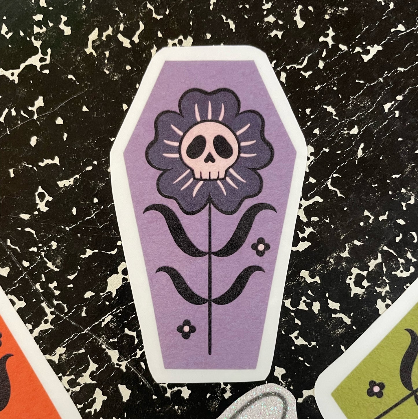 A coffin shaped sticker with a lavender gray background, featuring a black and gray flower with a skull in the center.