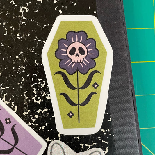 A coffin shaped sticker with a green background, featuring a black and gray flower with a skull in the center.