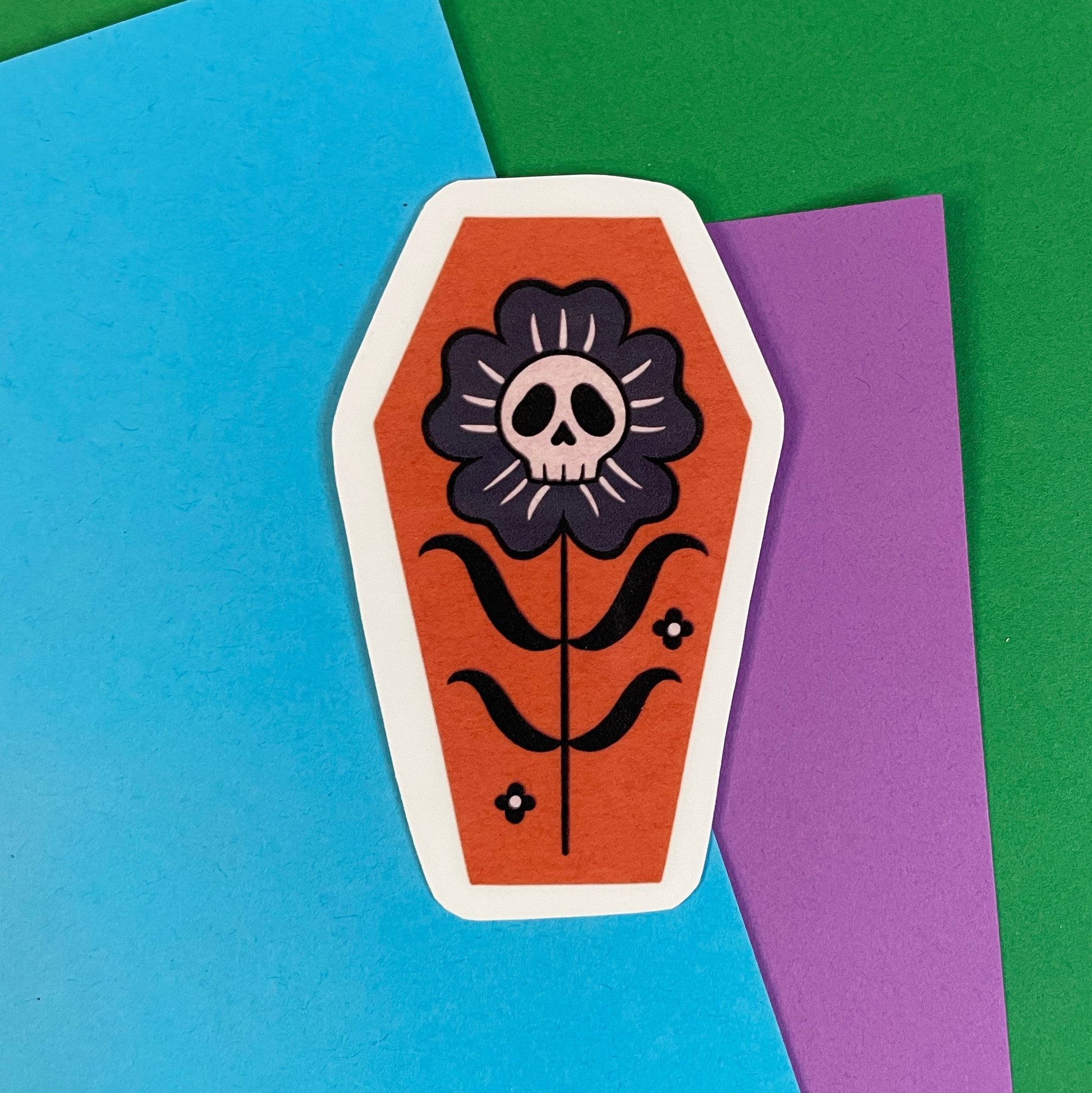 A coffin shaped sticker with an orange background, featuring a black and gray flower with a skull in the center.